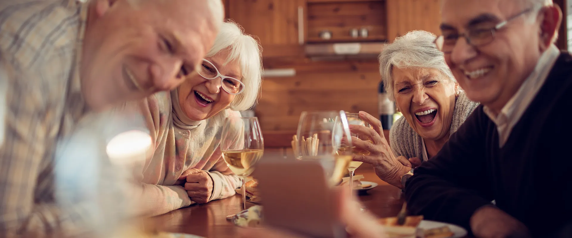 A heartwarming scene of four elderly friends gathered around a table, sharing laughter and enjoying a meal together in a cozy, wooden cabin setting. The joyful expressions highlight a moment of genuine connection and happiness.