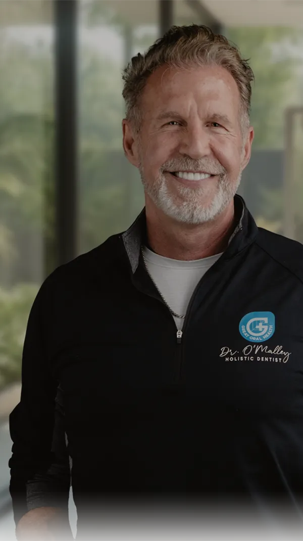 Dr. Paul O'Malley, dressed in a white lab coat, sits next to a reception desk with the word "SMILE" prominently displayed in large golden letters, beaming with a confident smile.