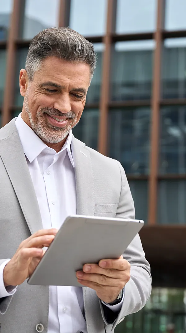 A middle-aged man smiling while looking at a tablet, standing outside a modern building, representing the confidence and satisfaction of a patient after receiving dental implants.