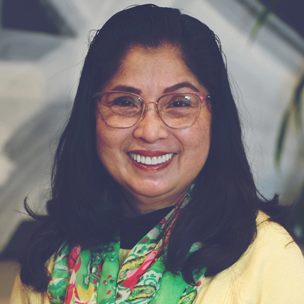 Close-up of Ynah Refuerzo, smiling warmly, wearing glasses and a colorful scarf over a yellow sweater, with a softly blurred indoor background.