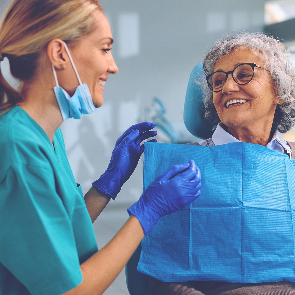 A female dental professional in teal scrubs cheerfully interacts with an elderly woman patient seated in a dental chair, both wearing protective masks. The dentist is adjusting a blue dental bib on the patient, preparing for a procedure.