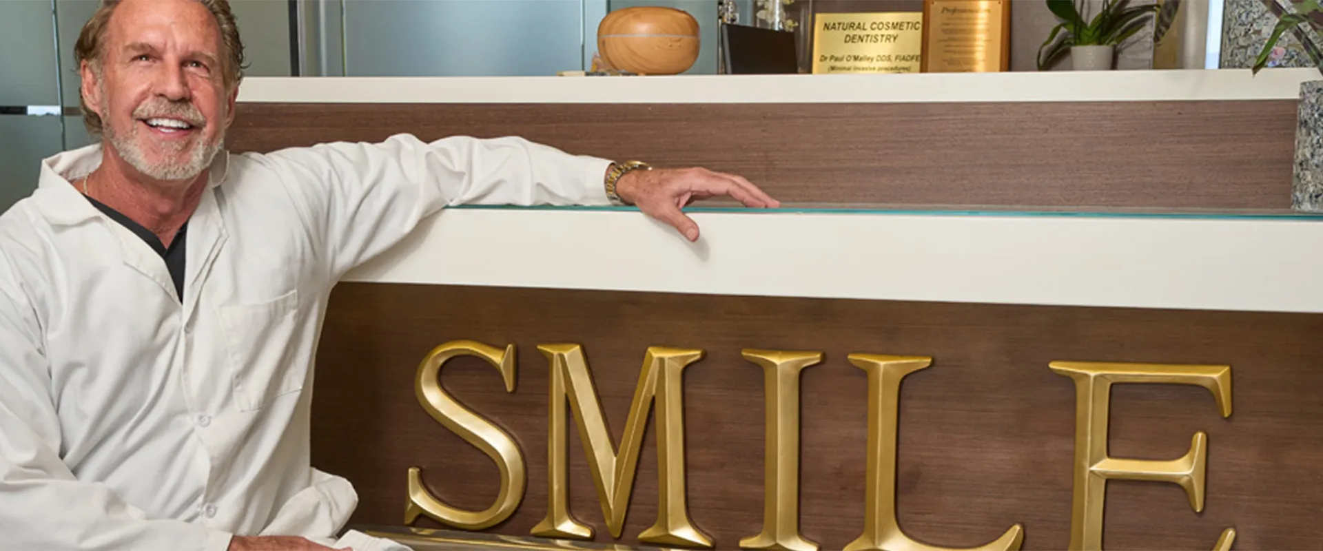 Dr. Paul O'Malley, dressed in a white lab coat, sits next to a reception desk with the word "SMILE" prominently displayed in large golden letters, beaming with a confident smile.