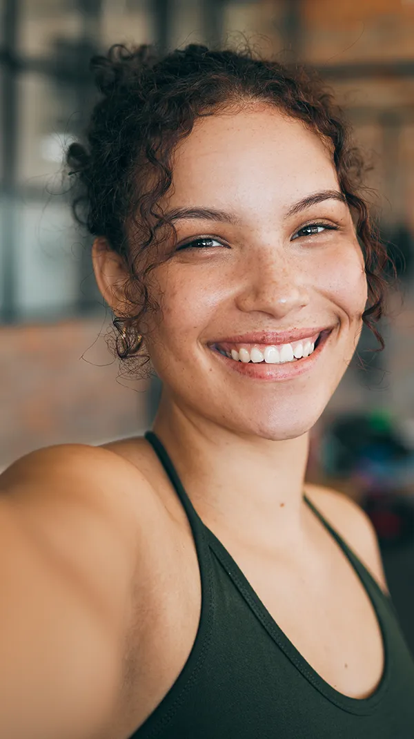 A young woman with curly hair and a bright smile in a gym setting, her healthy white teeth prominent, suggesting vitality and wellness