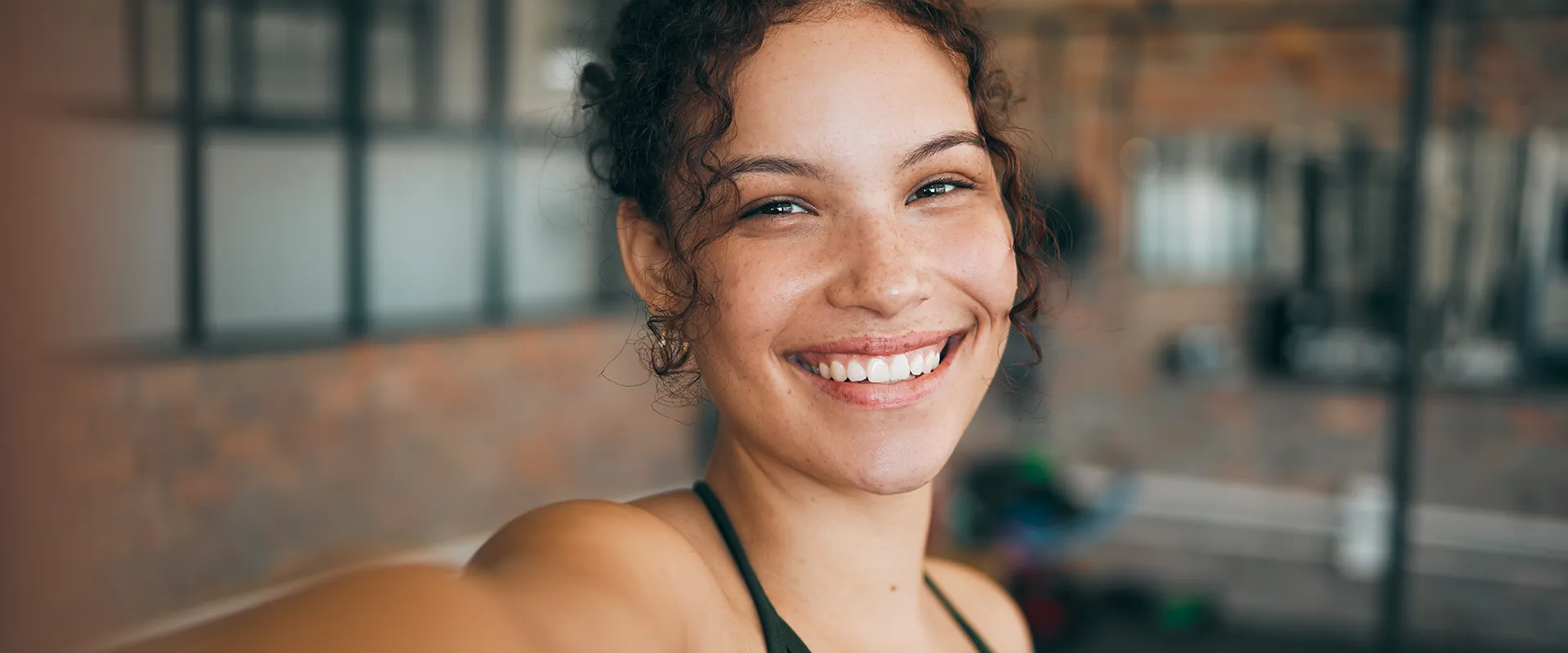 A young woman with curly hair and a bright smile in a gym setting, her healthy white teeth prominent, suggesting vitality and wellness