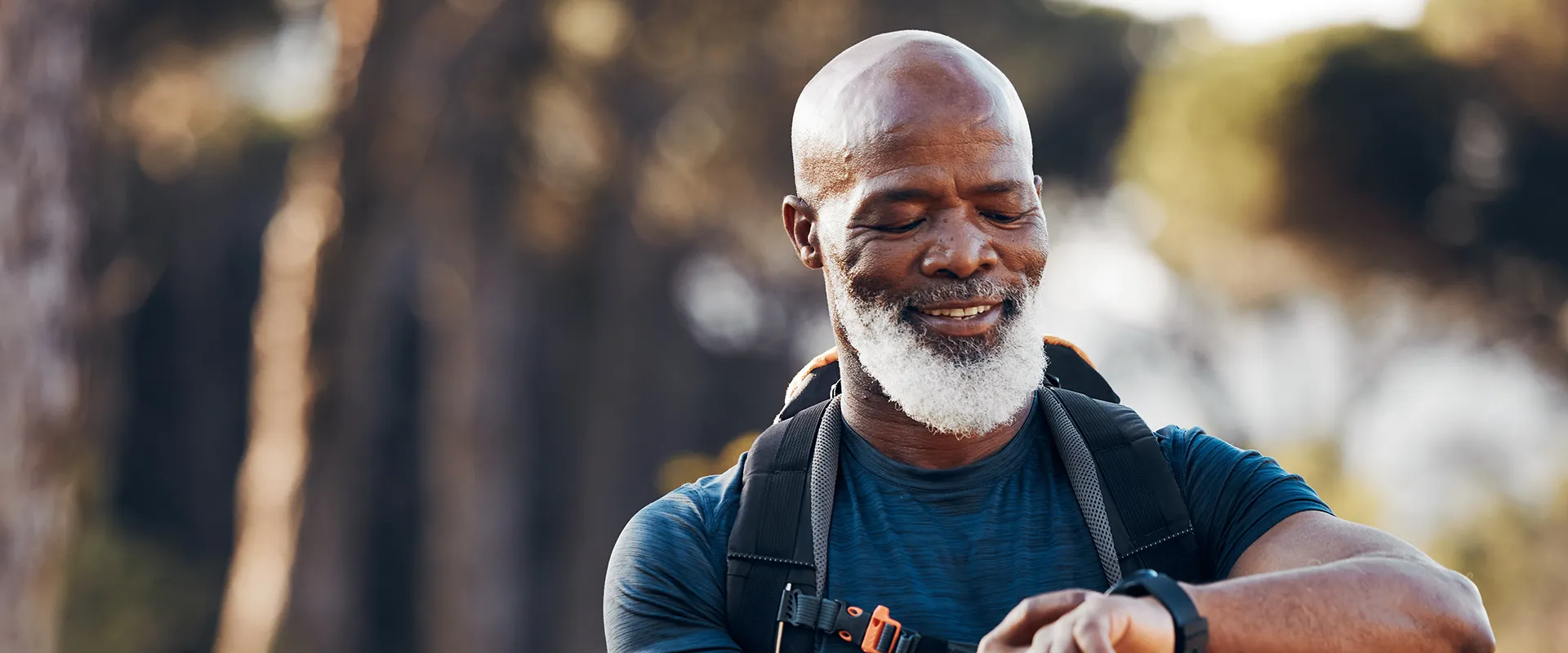 Smiling elderly man with a beard adjusting his watch while hiking, showcasing his vibrant and healthy smile in an outdoor setting, symbolizing an active lifestyle post full mouth reconstruction.