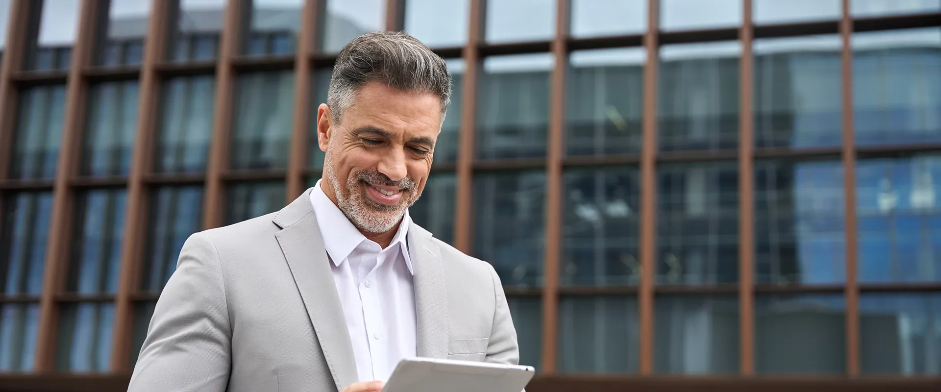 A middle-aged man smiling while looking at a tablet, standing outside a modern building, representing the confidence and satisfaction of a patient after receiving dental implants.