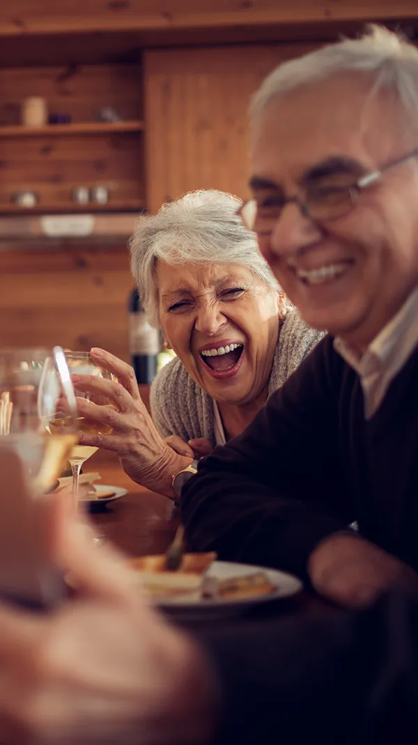 A heartwarming scene of four elderly friends gathered around a table, sharing laughter and enjoying a meal together in a cozy, wooden cabin setting. The joyful expressions highlight a moment of genuine connection and happiness.