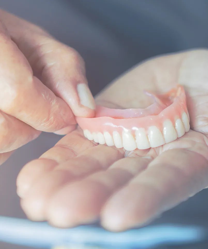 Close-up view of a pair of hands holding a set of upper dentures, demonstrating the realistic appearance and fit of the false teeth set against a gentle, blurred background.