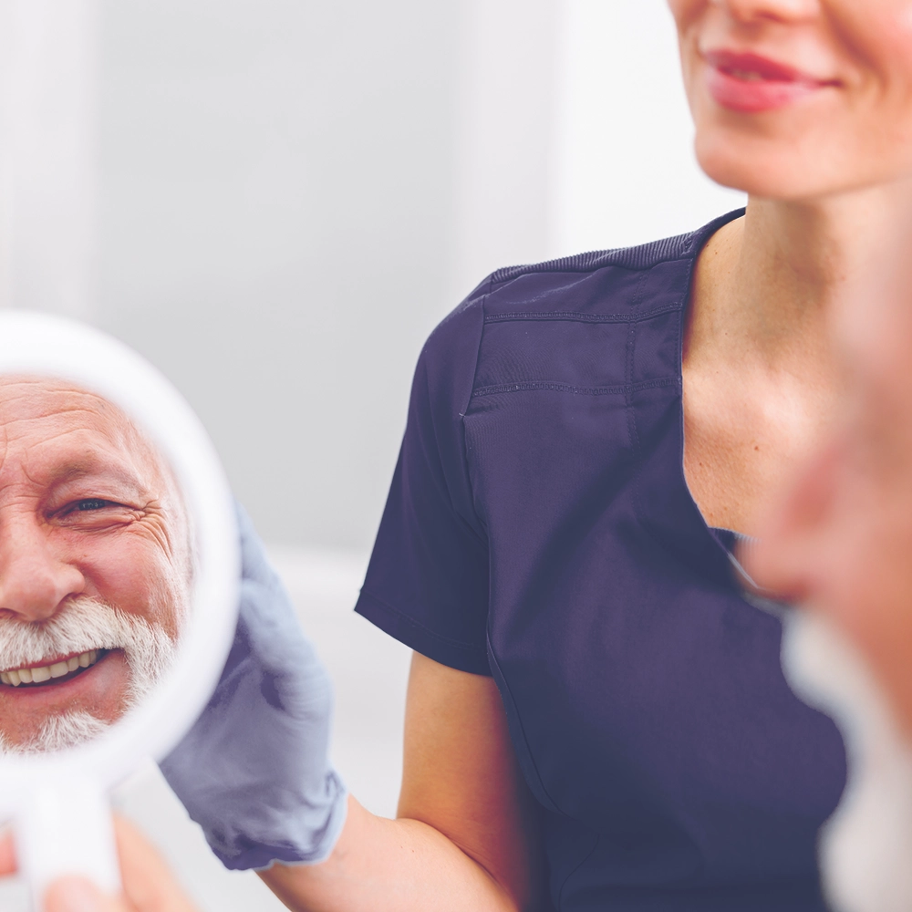 An elderly man smiles broadly while holding a mirror, examining his new dental work as a female dentist in navy blue scrubs looks on with a pleased expression, symbolizing successful dental care.