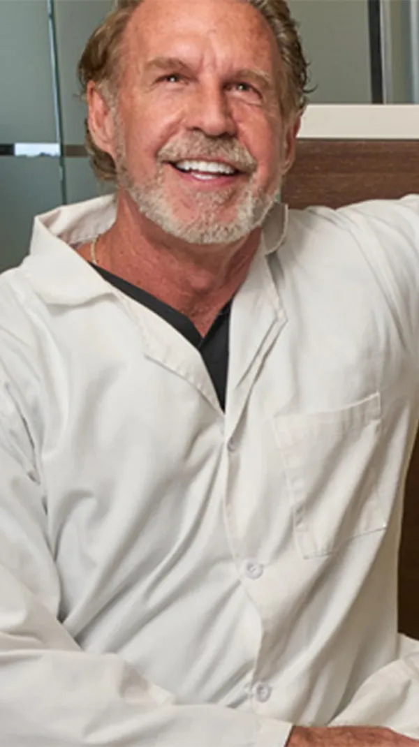 Dr. Paul O'Malley, dressed in a white lab coat, sits next to a reception desk with the word "SMILE" prominently displayed in large golden letters, beaming with a confident smile.