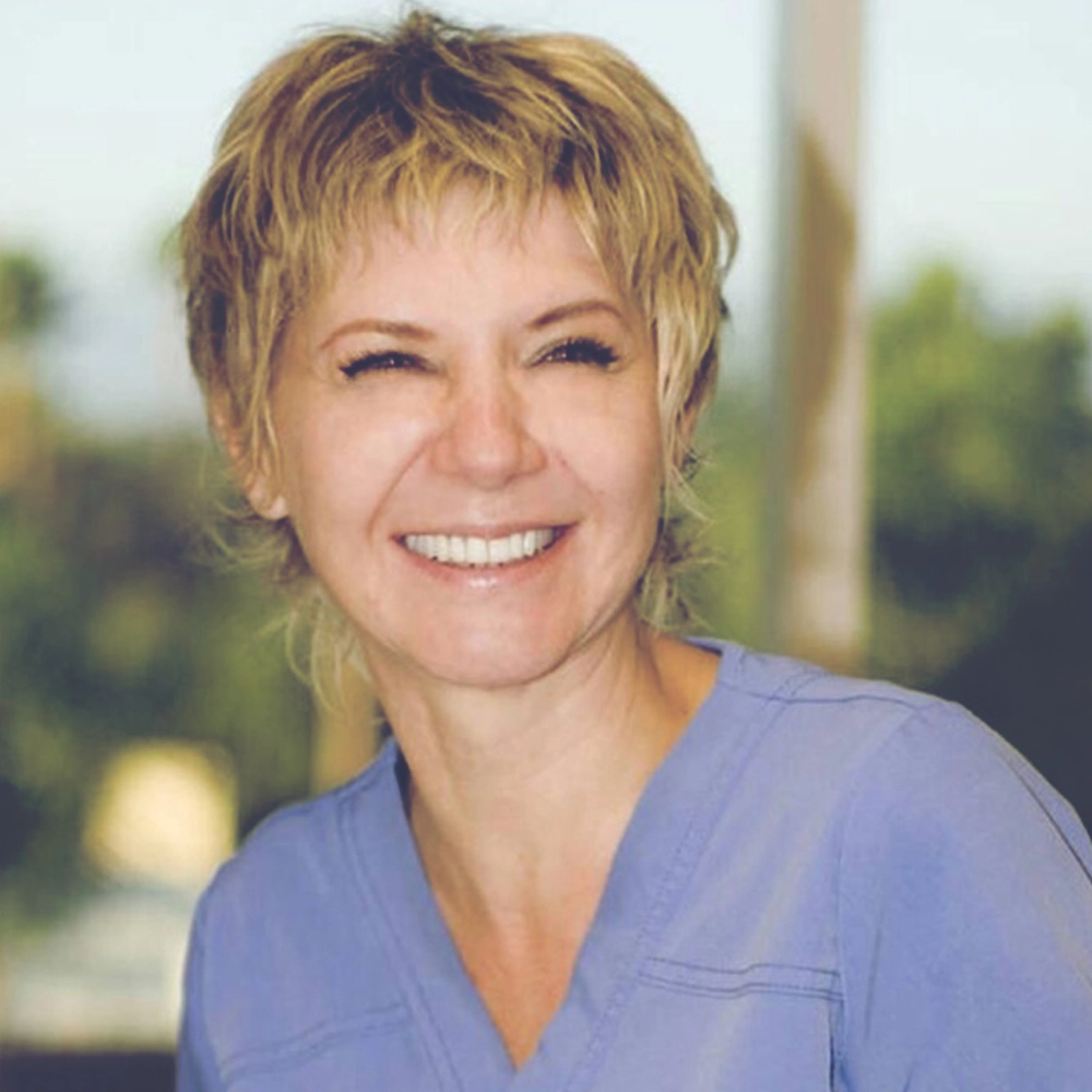 Portrait of Jenny Kozlova, smiling brightly while wearing a light blue scrub top, standing in front of a softly blurred natural background.