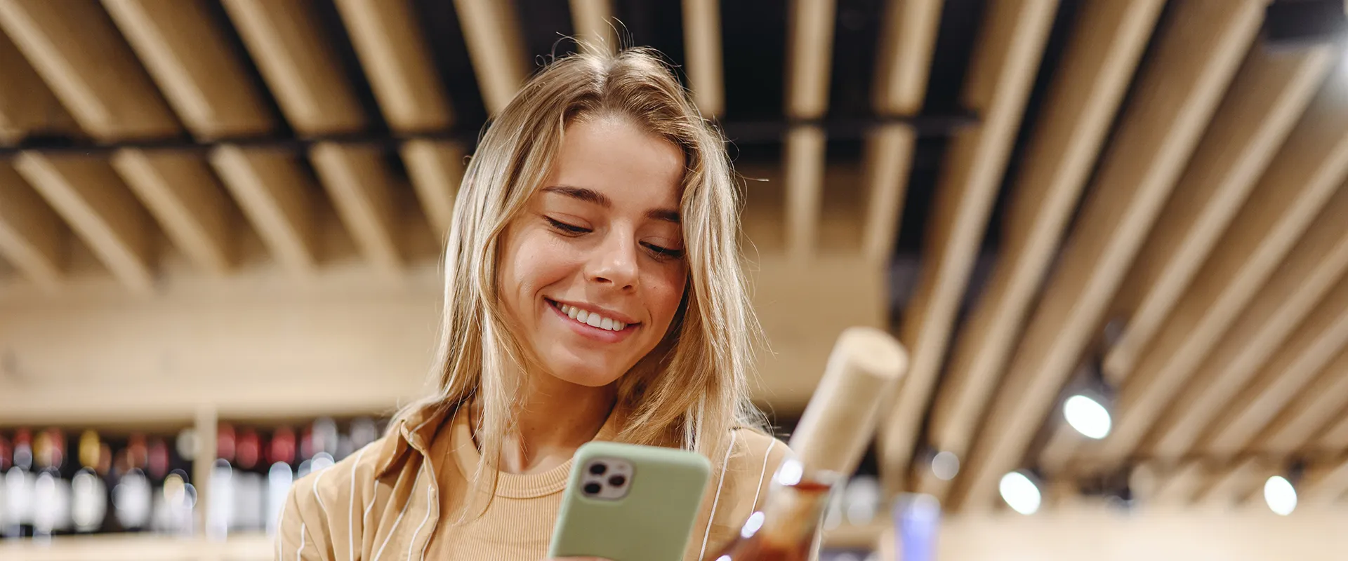 A joyful woman engaging with her smartphone, her smile showing healthy teeth, set against a contemporary backdrop with wooden beams overhead.